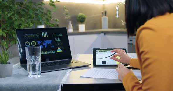 Woman in Stylish Jacket Sitting at Kitchen Table and Working with Documents alt