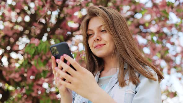 Smiling Woman Walks Down the Central Park City Street and Uses Her Phone alt