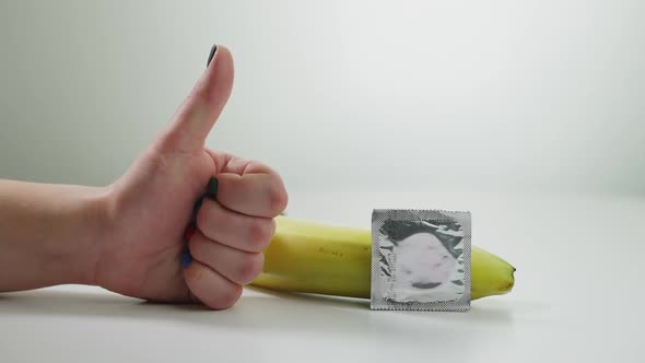 Closeup Female Hand Gesturing Thumb Up with Condom Lying at Banana alt