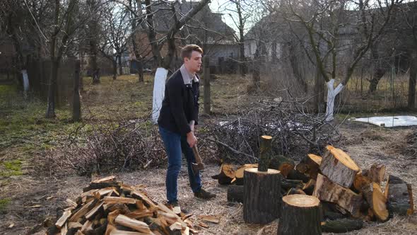 A young man is chopping wood with an axe in the garden near the greenhouse. camera movement