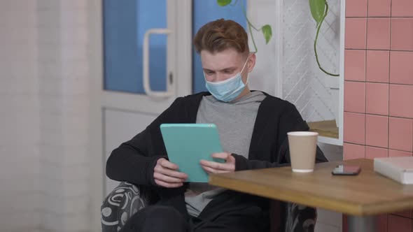 Portrait of Young Positive Man in Covid Face Mask Surfing Social Media on Tablet Smiling alt