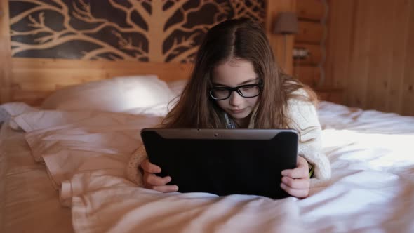 Cute Teenage Girl Browses the Internet on a Tablet Lying on a Bed in a Country Cozy House alt
