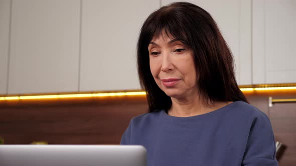 Smiling Aged Woman Uses Laptop Typing on Keyboard in Home Kitchen Distance Work alt