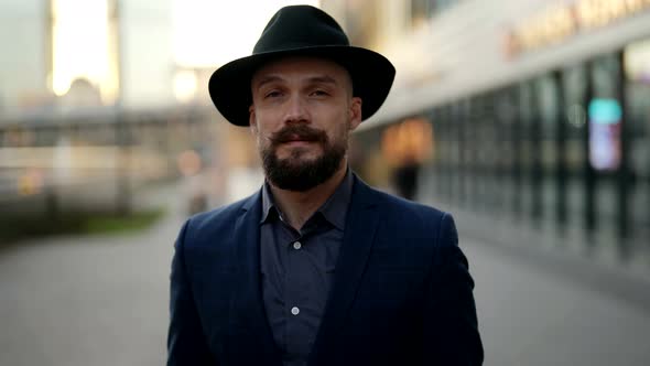 Portrait of a Bearded and Mustachioed Man in a Hat and a Blue Jacket at a Blurry Modern Building alt
