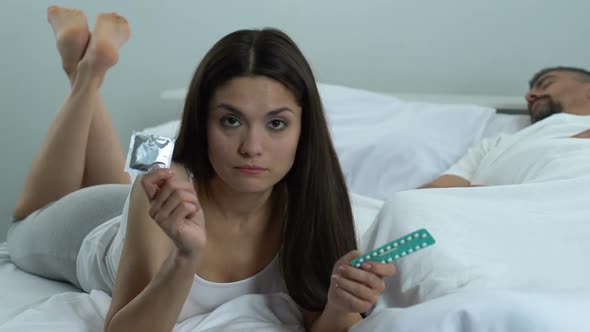 Puzzled Woman Holding Condom and Oral Pills, Contraception Choice, Health Care alt