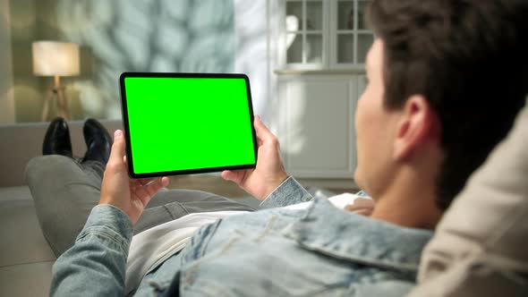 Over The Shoulder View Young Man at Home Lying on a Sofa Using With Green Mock-up Screen Tablet alt