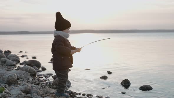 Baby Boy Playing with Stick and Water on the Stone Shore on an Autumn Evening alt
