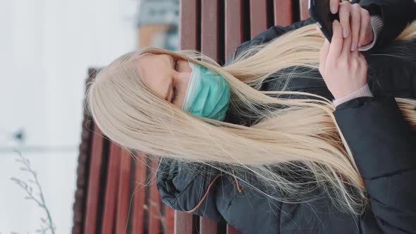 Young Female in a Medical Mask Sitting on the Bench and Using Smartphone alt