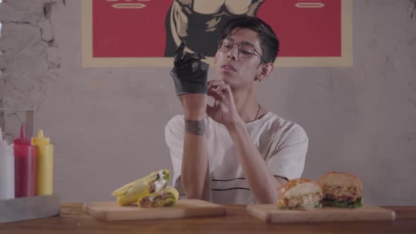 Bearded Man Sitting at the Table in Front of Mouth-watering Burger and Shawarma Preparing for Eating alt