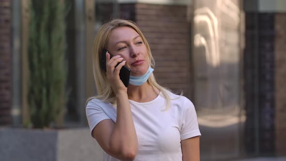 Portrait of Pretty Young Woman in Lowered Protective Medical Mask Is Talking on Her Smartphoneis alt