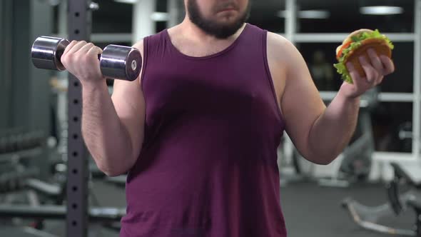 Overweight Male With Hamburger and Dumbbell in Hands Choosing Healthy Life alt