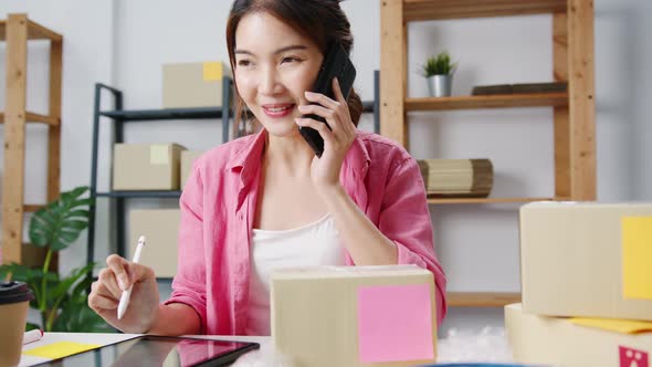 Young Asian woman using phone call receiving purchase order and check product on stock work at home. alt