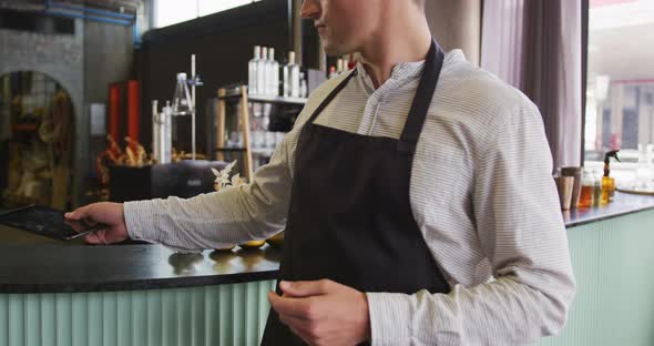 Portrait of caucasian man wearing apron working at a bar, using tablet and smiling to camera alt