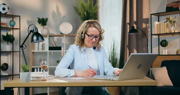 Woman in Headset Working with Documents and Information on Laptop at Workplace in Cozy Room alt