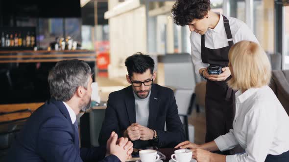Bearded Man in Suit Making Online Payment in Cafe Then Talking To Coworkers alt