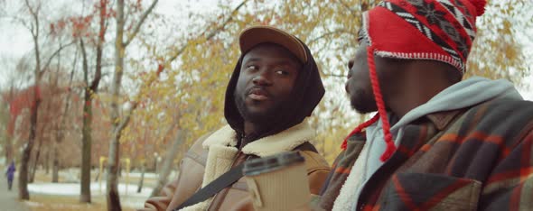 African American Friends Drinking Coffee and Chatting on Bench in Park alt