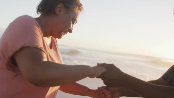 Smiling senior african american couple holding hands and dancing on sunny beach alt