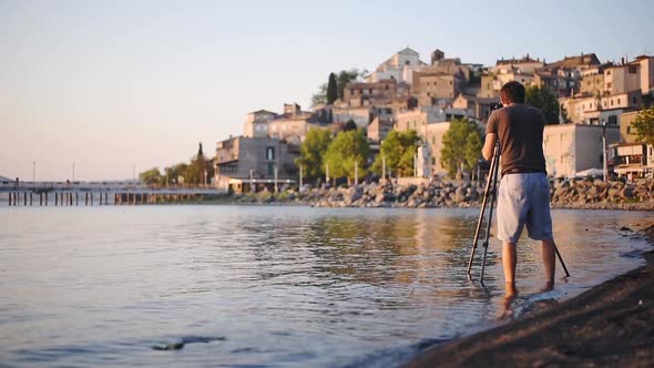 A Man Wearing Shorts Standing In The Lake Bracciano Shore With His Camera Capturing The Beauty Of Ca alt