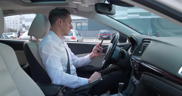 Smiling Male Entrepreneur Sits in the Driver's Seat in a Car and Uses a Modern Smartphone