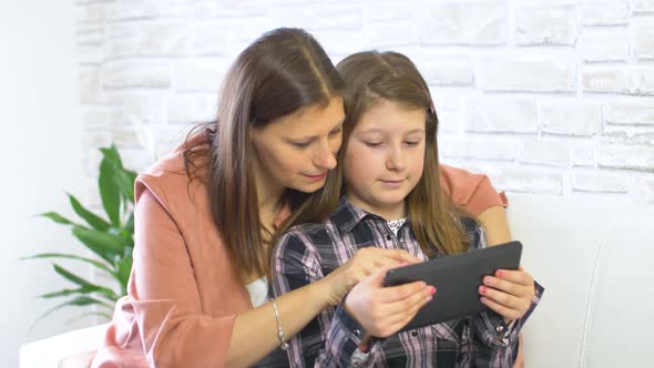 Happy mother and her daughter smiling having fun using digital tablet sitting on white sofa at home alt