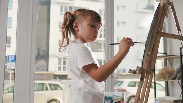 Pretty Little Girl Paints with Brush on Canvas on an Easel
