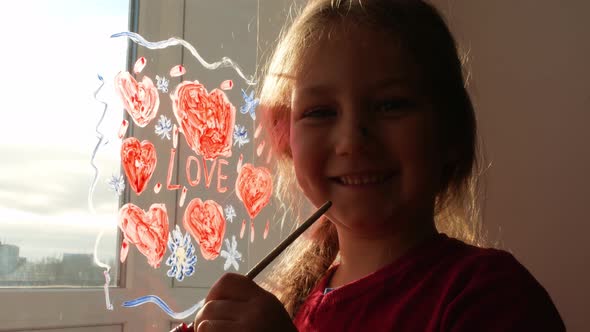 Little girl holds paintbrush in hand draws red heart on window Valentine's day love home family art