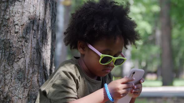 A Little Black Girl in Plastic Shades Sitting Under the Tree in the Park and Playing a Game on the alt