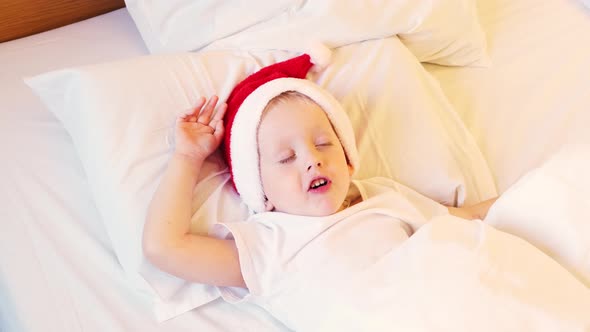 Funny christmas. Little boy in Santa hat lying in white bed and pretends to be asleep. alt