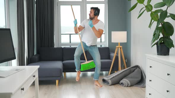 Man Listens to Music with Headphones and Cleaning the House and Having Fun Dancing with a Broom alt