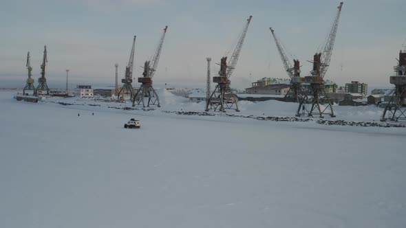 A White SUV Drives Down a Frozen River in Winter Past Port Cranes in the Arctic