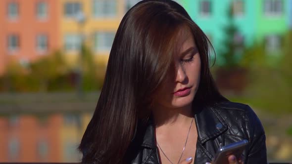 Young Attractive Girl, Prints a Message on a Smartphone in the Street in the Park