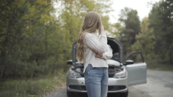 Charming Slim Young Woman Talking on the Phone As Man Repairing Car Engine at the Background alt