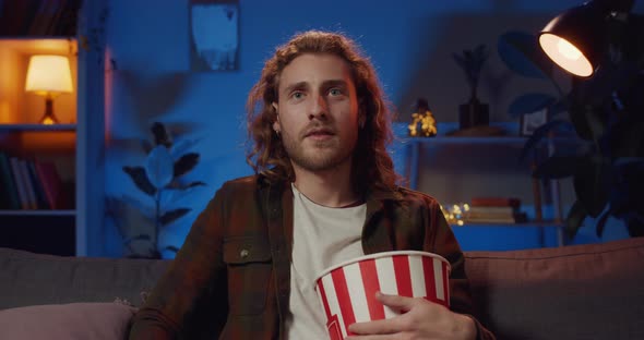 Good Looking Young Man Holding Popcorn Paper Bowl and Eating While Switching on Tv, Cheerful Hipster