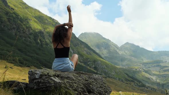 A cinematic view of a beautiful young woman sitting on a rock and enjoying the view of the mountain alt