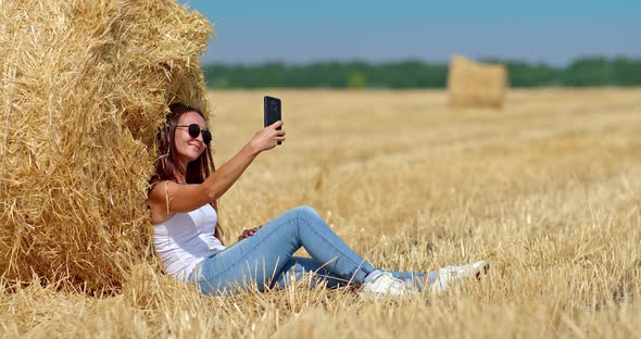 Portrait a Girl in Sunglasses is Sitting By a Large Haystack and Taking a Selfie on a Mobile Phone alt