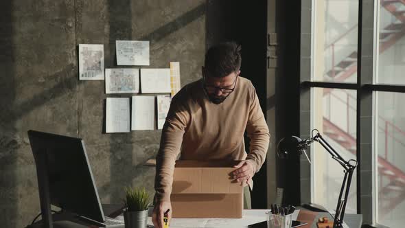 Young Man Unpacks a Cardboard Box in New Office and Takes Out a Plant