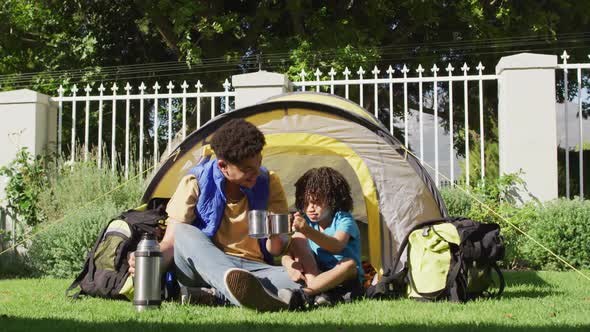 Happy biracial man and his son drinking from thermos flask in garden alt