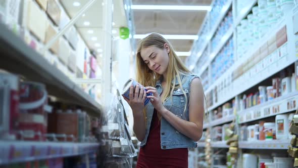 Young Female Customer is Taking Two Jar with Paint in a Store for Builders alt