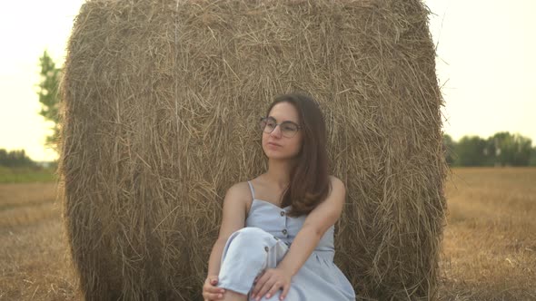 A Young Woman Sits Near a Haystack in a Field alt