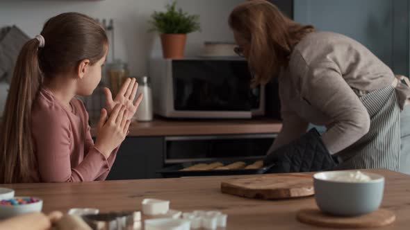 Grandmother and granddaughter taking ready cookies out of the oven. alt