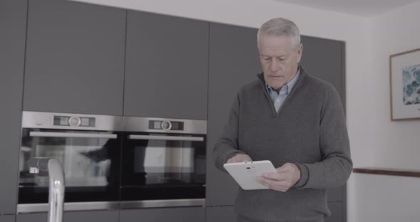 Senior adult man in kitchen checking email on digital tablet at home in casual clothes