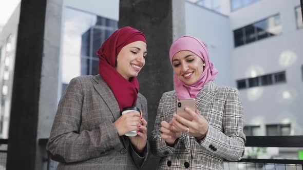 Two Young Muslim Women Wearing Hijab Headscarf Look at Phone Walking Together Laughing Smile To Eash