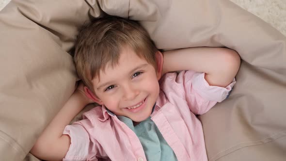 Camera Flying Away From Happy Smiling Boy Lying in Beanbag Chair in Living Room alt