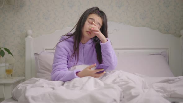 Sleepy 20s Asian Woman Browsing Internet Using Cell Phone Sitting in Bed in the Evening