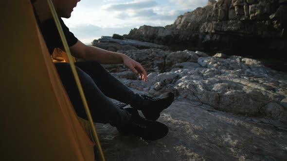 Young man in a black T-shirt is resting in a tent on the rocks by the sea alt
