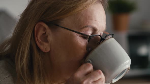 Extreme close up of thoughtful caucasian senior woman sitting in armchair, drinking coffee alt