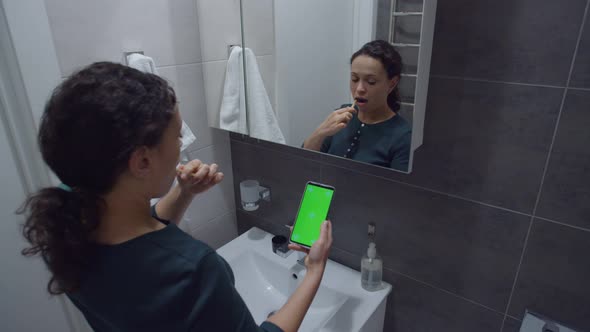 Black Woman Looking at Smartphone with Chroma Key While Brushing Teeth alt