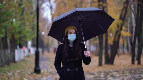 A Young Woman in a Protective Mask Walking in the Park Under Umbrella. Rainy Day, During Second Wave