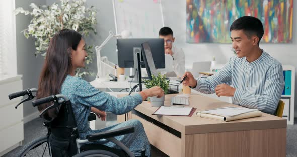 Woman is Sitting in Wheelchair at Desk in Office Across Employer They are Signing Employee Contract alt