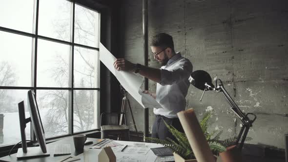 Bearded Man Standing Faces To Panoramic Window and Looks at Blueprints